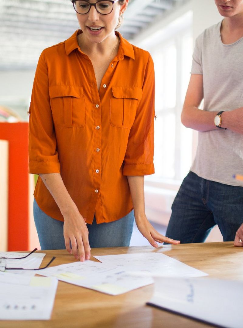 Bespectacled woman in an orange shirt and a man in a T-shirt and jeans stand over a desk scattered with papers and a pair of glasses.